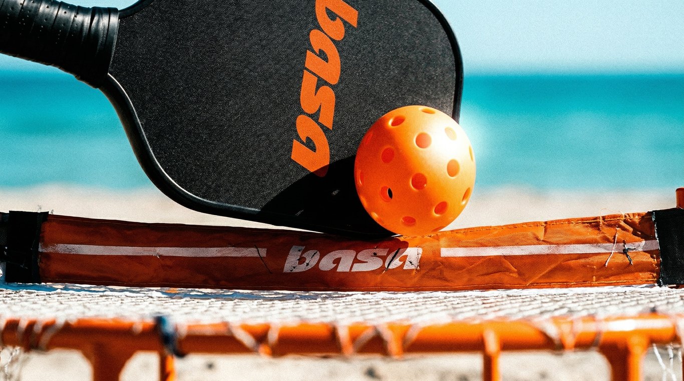 Close-up of a Basa paddle and orange pickleball resting on a portable net at the beach